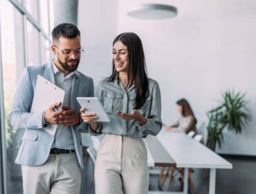 Shot of a two confident business persons talking in the work place. Two colleagues using a digital tablet while walking in a modern office. Businessman and businesswoman in meeting discussing business strategy. Business coworkers working together in the offic